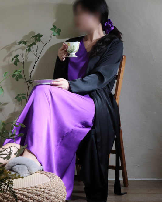 A woman elegantly seated, wearing a Royal Purple mulberry silk maxi dress paired with a long black robe. She holds a teacup, exuding a sense of calm and sophisticated comfort, highlighted by the matching royal purple silk scrunchie in her hair. This image showcases the versatile and luxurious styling potential for both relaxed moments and refined everyday wear.