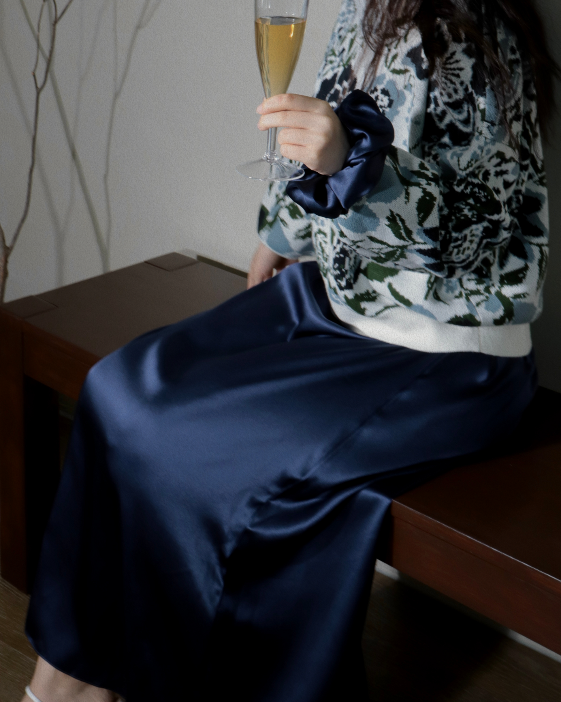 Woman sitting, holding a champagne glass, elegantly wearing a Midnight Navy 100% mulberry silk maxi dress. The image highlights the luxurious drape and flow of the maxi dress, perfect for special occasions or sophisticated loungewear. A matching silk scrunchie is visible on her wrist.