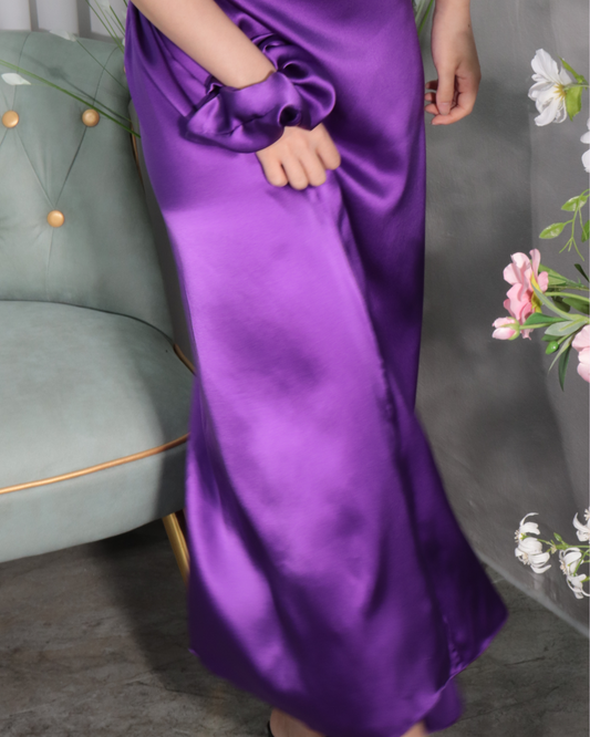 Close-up shot of a woman's lower body, showcasing the elegant drape and smooth texture of the Royal Purple mulberry silk maxi dress. The image highlights the fluid movement of the fabric as she subtly shifts, with a matching royal purple scrunchie visible on her wrist, emphasizing the luxurious feel and natural flow of the silk.