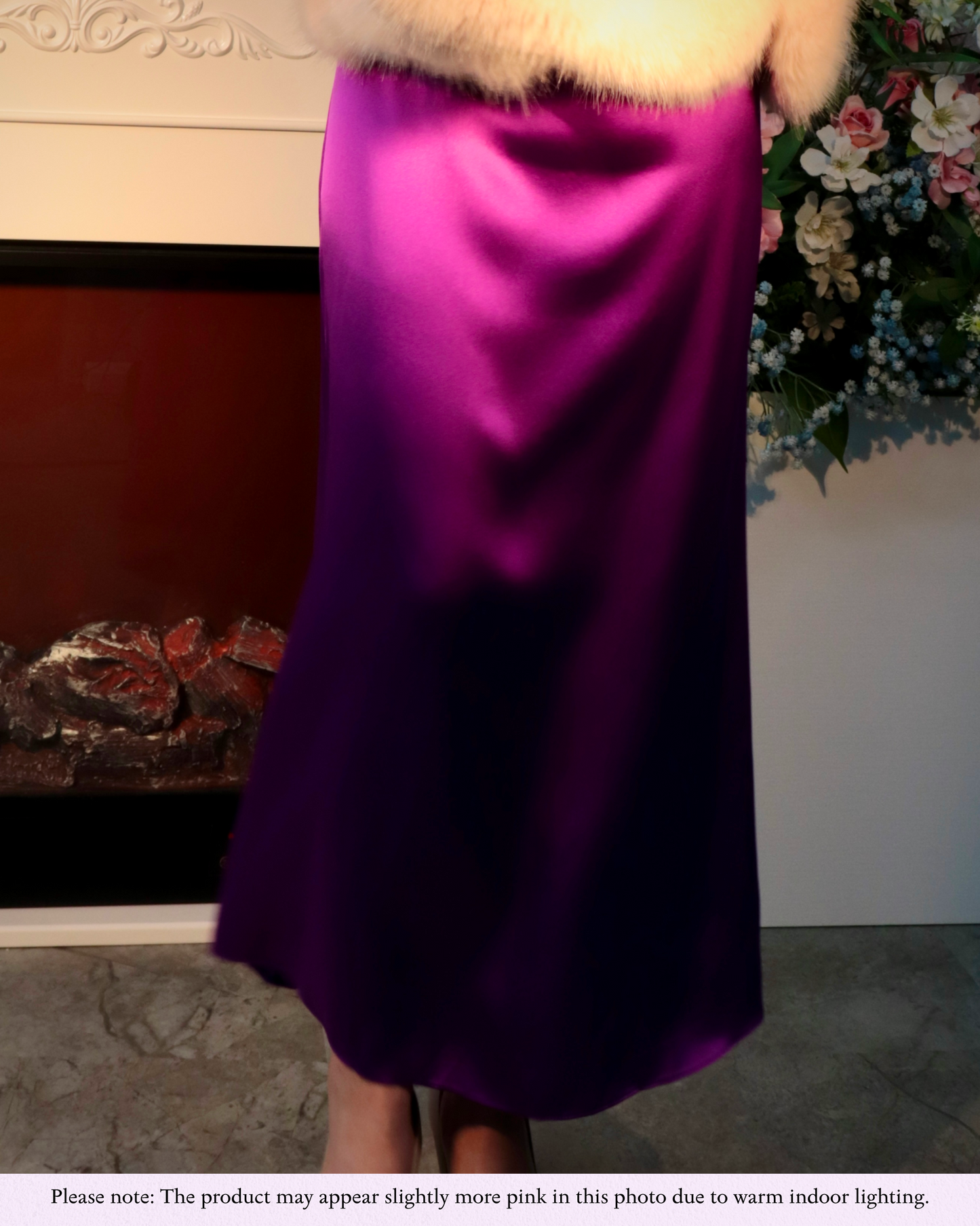 Close-up shot focusing on the lower half of a woman, showcasing the Royal Purple mulberry silk maxi dress in a festive setting with a fluffy jacket. The image highlights the dress's luxurious drape and smooth texture, with soft lighting creating a warm ambiance. A note at the bottom clarifies that the product may appear slightly more pink in this photo due to warm indoor lighting, ensuring accurate color representation despite the lighting conditions.