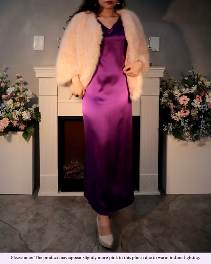 A woman elegantly posing in a Royal Purple mulberry silk maxi dress, styled with a light, fluffy jacket, in a setting reminiscent of a festive occasion. She holds her hands near her waist, showcasing the dress's luxurious drape and rich color. A note at the bottom clarifies that the product may appear slightly more pink in this photo due to warm indoor lighting, ensuring accurate color representation despite the lighting conditions.