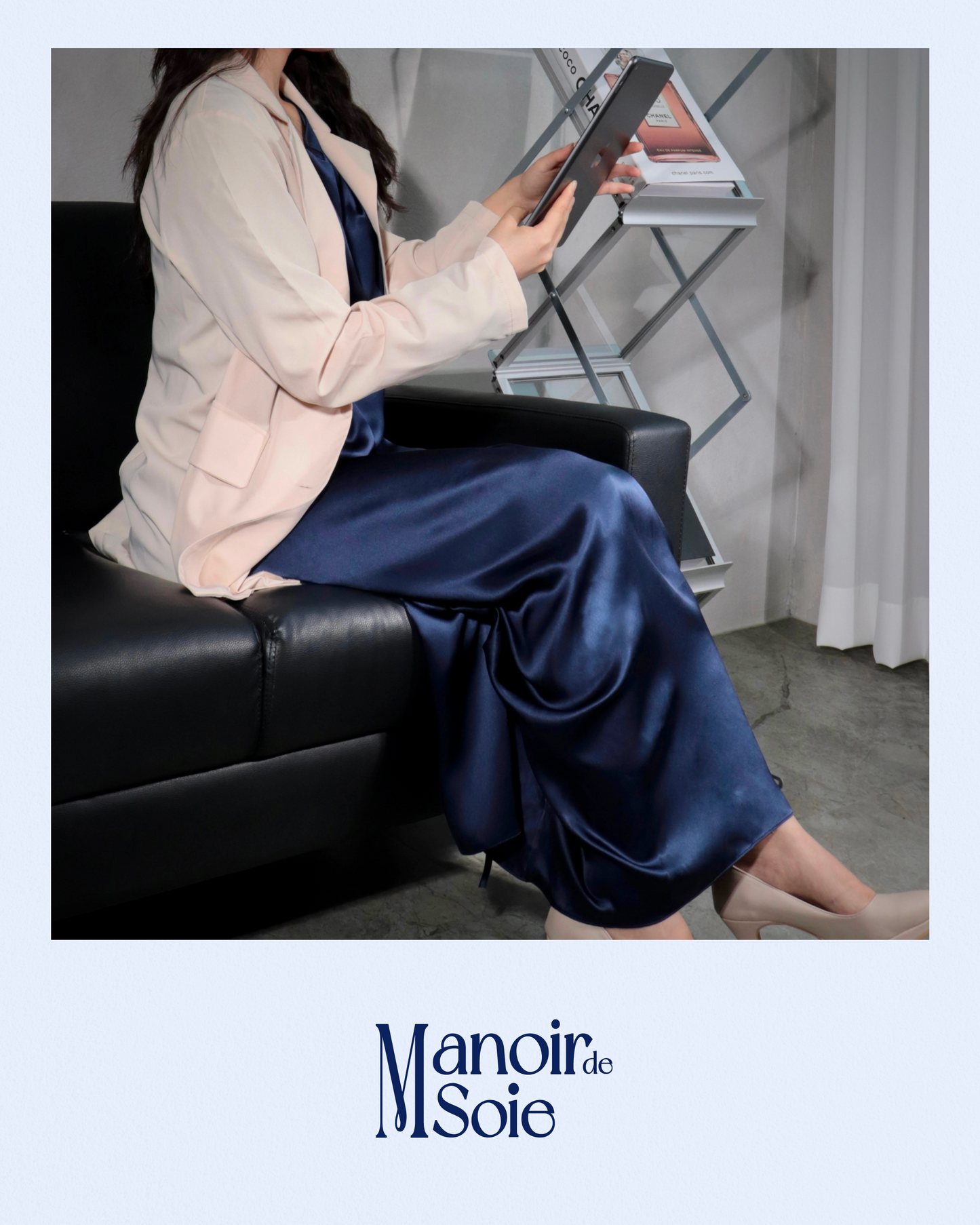 Woman sitting gracefully on a black sofa, wearing a Midnight Navy mulberry silk dress layered with a light blazer, reviewing a tablet. This image highlights the versatility and sophistication of the silk dress, suitable for both professional settings and elegant casual wear. Features "Manoir de Soie" logo.