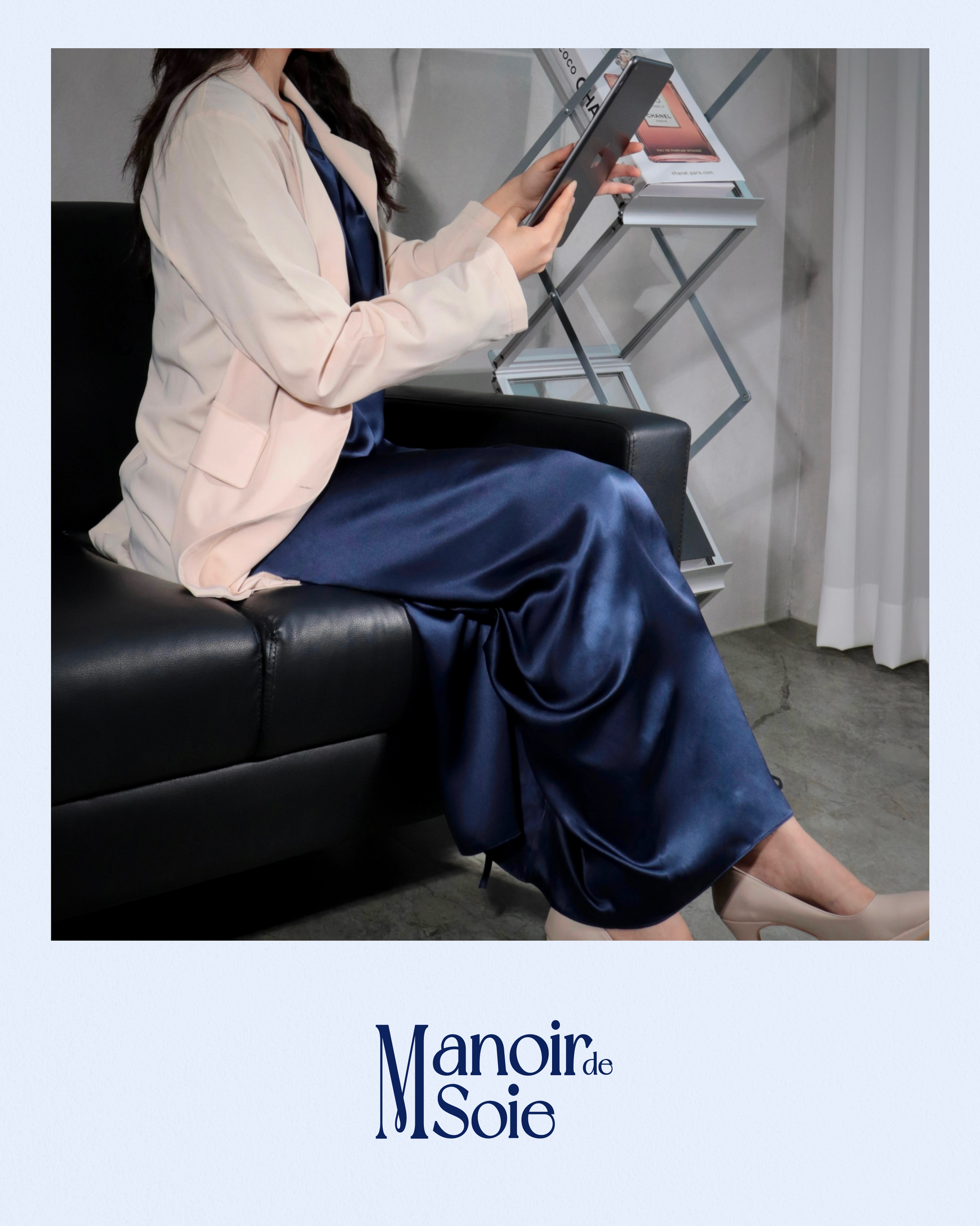 Woman sitting gracefully on a black sofa, wearing a Midnight Navy mulberry silk dress layered with a light blazer, reviewing a tablet. This image highlights the versatility and sophistication of the silk dress, suitable for both professional settings and elegant casual wear. Features "Manoir de Soie" logo.