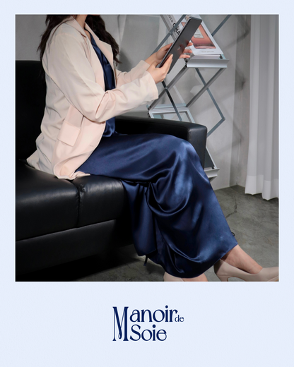 Woman sitting gracefully on a black sofa, wearing a Midnight Navy mulberry silk dress layered with a light blazer, reviewing a tablet. This image highlights the versatility and sophistication of the silk dress, suitable for both professional settings and elegant casual wear. Features "Manoir de Soie" logo.
